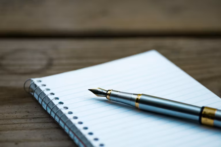 A fountain pen resting on an open spiral notebook on a wooden table
